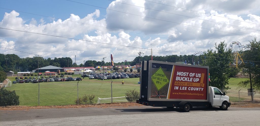 Picture of a moving billboard truck with Most of Us Buckle Up In Lee County ad parked on road in front of a fair in Lee County.