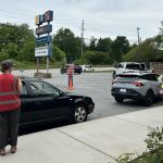 People in safety vests direct cars in a parking lot near a sign for a business, with trees and power lines in the background.