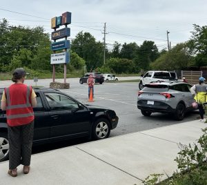 People in safety vests direct cars in a parking lot near a sign for a business, with trees and power lines in the background.