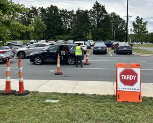A person in a safety vest stands by a car in a parking lot near a sign that reads, TARDY It is now after 8:15 AM. Orange safety cones are placed nearby.