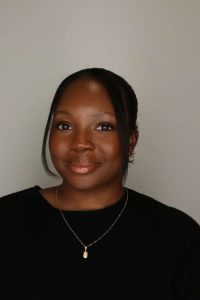 A headshot of a smiling person wearing a black shirt and a gold necklace.