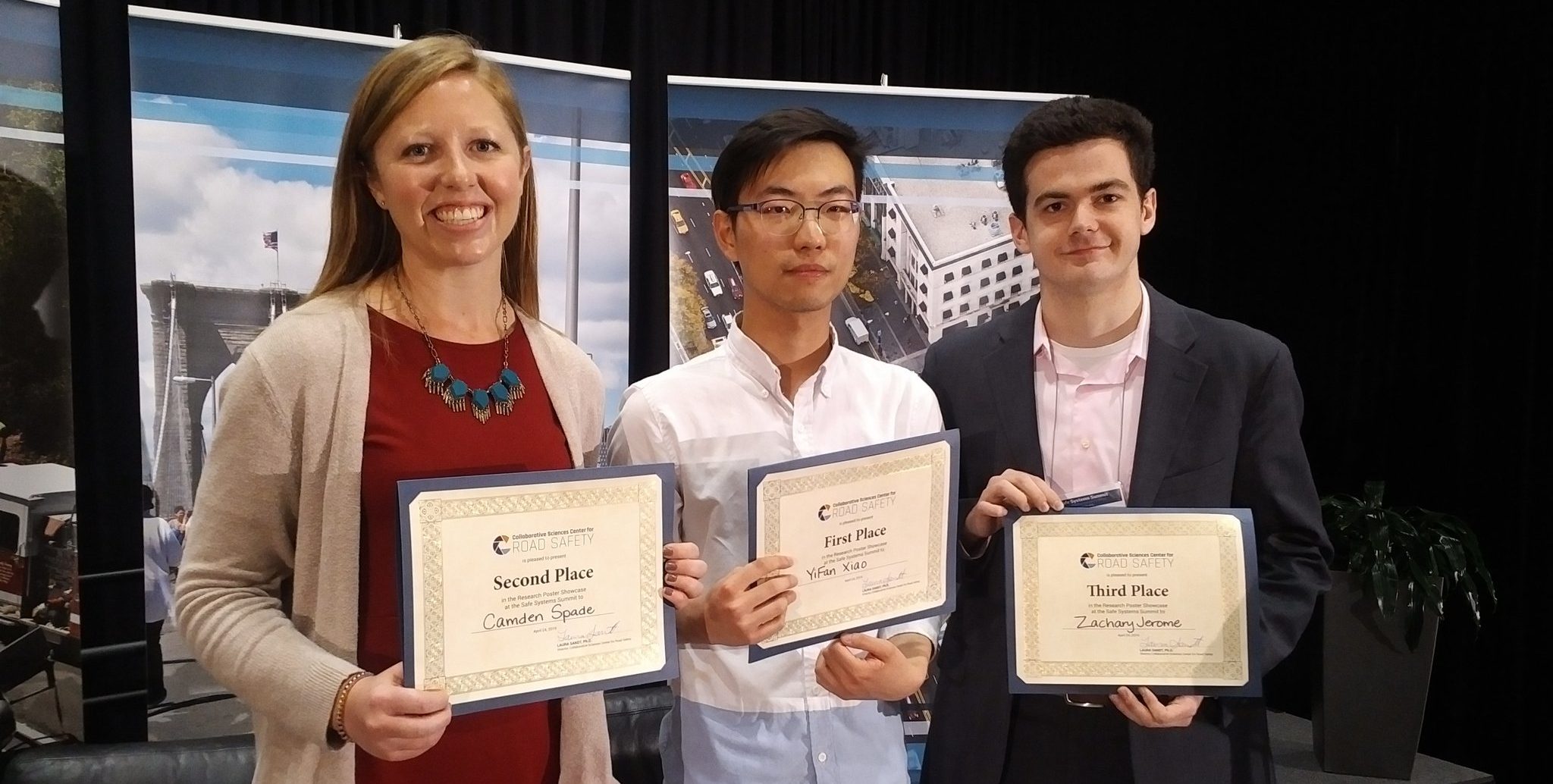 Three people stand indoors holding certificates for first, second, and third place awards. They are posing in front of a stage backdrop and smiling at the camera.