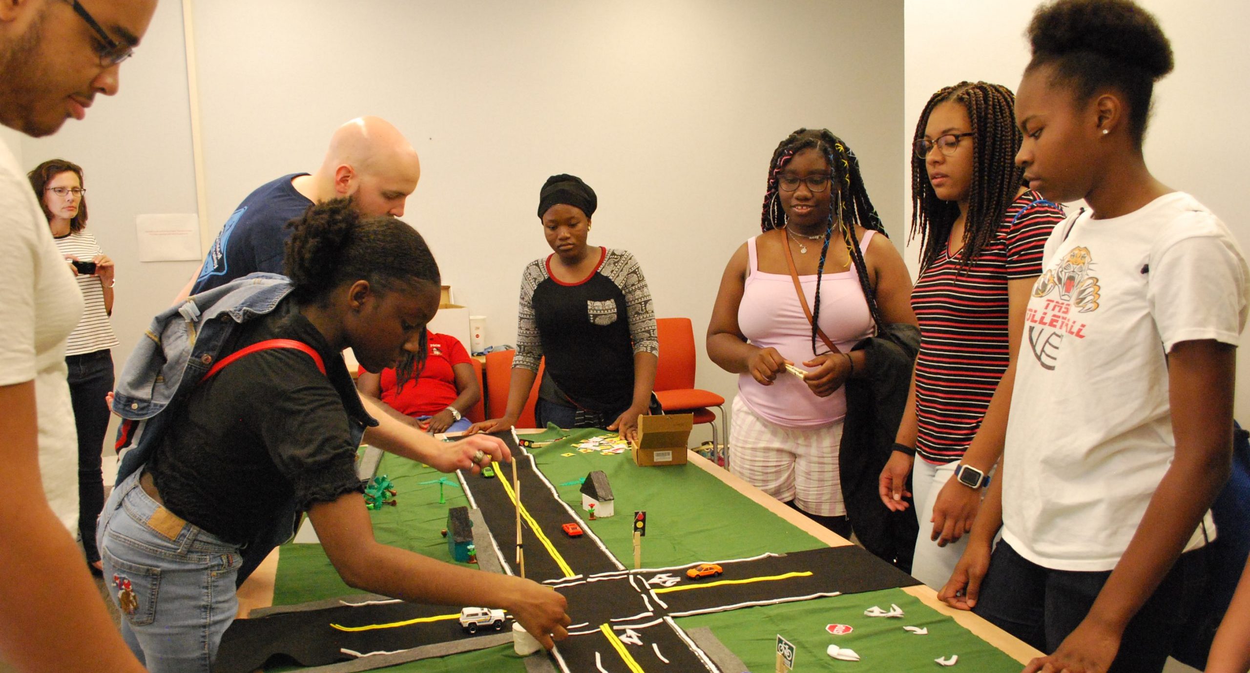 A group of people stand around a table with a model of intersecting roads, placing small objects and cars on the setup.