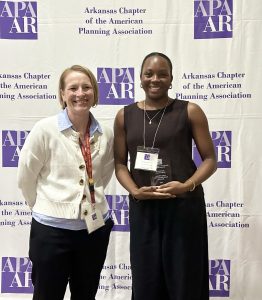 Two women standing in front of an Arkansas Chapter of the American Planning Association step-and-repeat banner, with one woman holding an award.