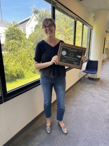 A woman stands indoors by a large window, smiling and holding a plaque from the University of North Carolina at Chapel Hill School of Social Work.
