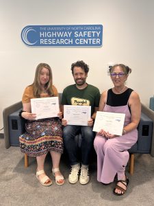 Three people sit on a couch holding certificates at the University of North Carolina Highway Safety Research Center, with the centers sign on the wall behind them.